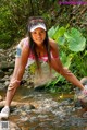 A woman in a pink top and denim shorts standing on a rock in a stream.