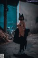 A woman wearing a black cat mask standing in an alleyway.